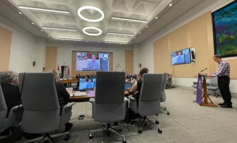 Board room, people sitting at a round table with large office chairs, man standing at a podium speaking.