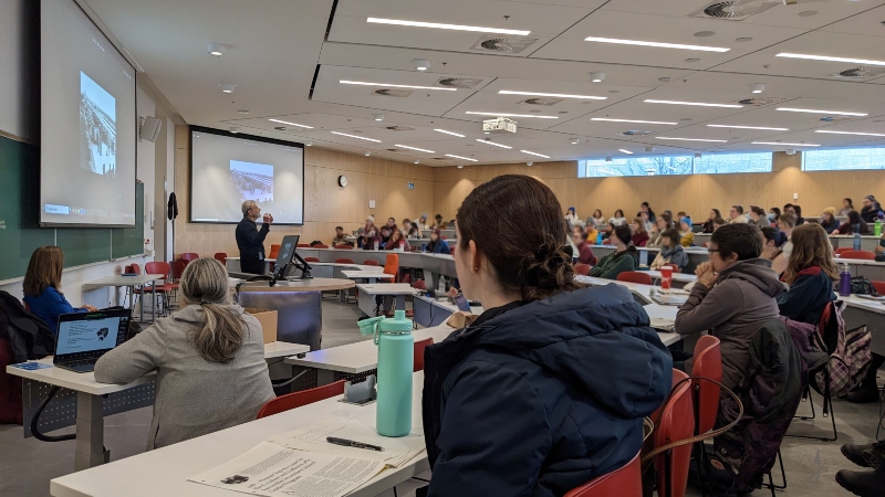Students in a University lecture hall sitting at rows of white table with red chairs that form a semi-circle around the professor. He is speaking with 2 screens behind him with the same image projected on each screen.
