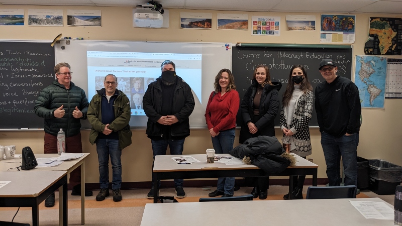 7 teachers 4 male, 3 female, standing, behind them a chalkboard and Whiteboard with the CHES presentation slide projected on it. 